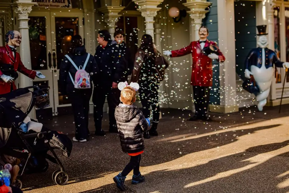 A child wearing mouse ears enjoys bubbles at an amusement park entrance.