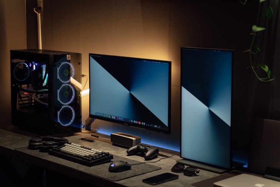 Modern dual monitor PC setup with tech gadgets on a sleek desk in a dimly lit room.